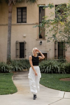 full length of pleased young woman in white cargo pants and trendy tank top standing near house in Miami