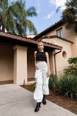 In voller Länge steht eine junge Frau in trendigen Klamotten und Stiefeln in der Nähe moderner Häuser und grüner Pflanzen in Miami 