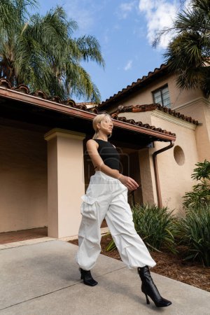 volle Länge der jungen Frau in trendigen Kleidern zu Fuß in trendigen Stiefeln in der Nähe von modernen Haus in Miami 