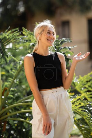 Foto de Alegre joven mujer en negro tanque superior y pantalones blancos de pie cerca de plantas verdes - Imagen libre de derechos