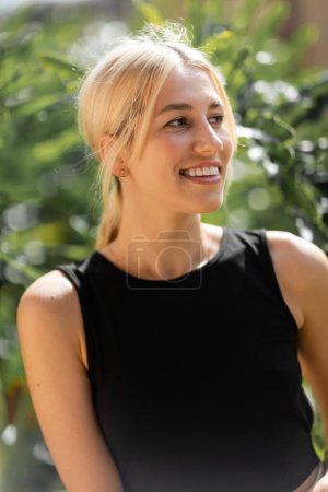 portrait of cheerful young woman in black tank top smiling near green plants 