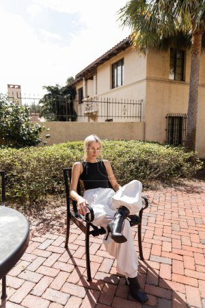 in voller Länge junge Frau in trendigen Klamotten sitzt auf Terrassenstuhl in der Nähe von modernem Haus in Miami 