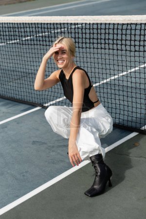 full length of happy woman in tank top and white cargo pants sitting near net in tennis court