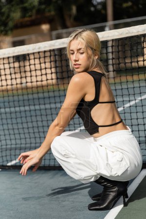 full length of young woman in tank top and white cargo pants sitting near net in tennis court
