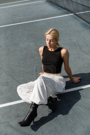 high angle view of young woman in tank top and white cargo pants sitting on tennis court