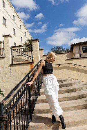 Téléchargez les photos : Pleine longueur de femme blonde en débardeur et pantalon cargo blanc debout sur les escaliers près du bâtiment à Miami - en image libre de droit
