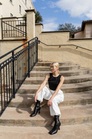 Blonde Frau in angesagtem Outfit sitzt auf einer Treppe in der Nähe eines Gebäudes in Miami 