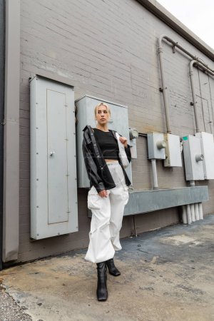 full length of young blonde woman in leather shirt jacket and cargo pants standing with handbag on industrial street in Miami 