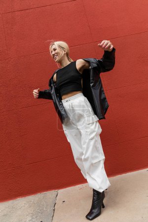 full length of cheerful woman in cargo pants and leather shirt jacket standing near red wall in Miami 