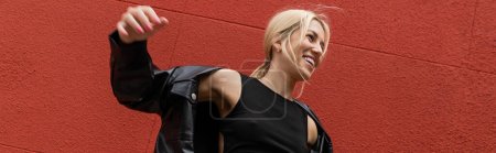 Photo for Cheerful woman in cargo pants and leather shirt jacket standing near red wall in Miami, banner - Royalty Free Image
