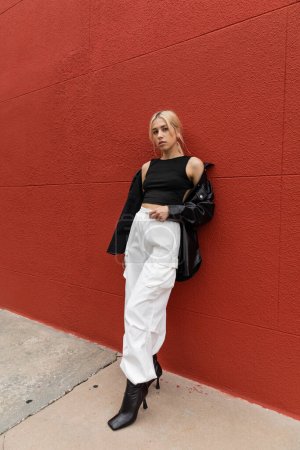 full length of blonde woman in cargo pants and leather shirt jacket standing near red wall in Miami 
