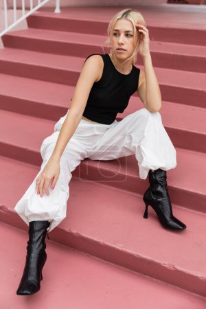 Téléchargez les photos : Pleine longueur de jeune femme blonde en débardeur noir et pantalon cargo assis sur des escaliers roses à Miami - en image libre de droit