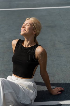 Téléchargez les photos : Femme gaie en débardeur noir assis sur le court de tennis à l'extérieur - en image libre de droit