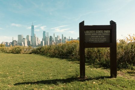 Foto de NEW YORK, USA - OCTOBER 13, 2022: board with liberty state park lettering on green lawn and cityscape with skyscrapers on background - Imagen libre de derechos