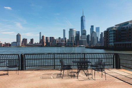 terraplén con mesa y sillas cerca del puerto y paisaje urbano de Manhattan en la ciudad de Nueva York