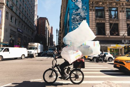 Foto de NEW YORK, USA - OCTOBER 13, 2022: delivery man with cellophane bags riding bicycle on crossroads of city street - Imagen libre de derechos