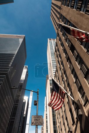 Téléchargez les photos : Vue à angle bas des bâtiments avec des drapeaux américains contre le ciel bleu dans le quartier de Manhattan à New York - en image libre de droit