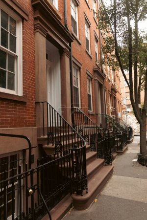 Backsteingebäude mit weißen Fenstern und Eingängen mit Treppen und Metallzäunen in der Nähe eines Baumes auf einer städtischen Straße in New York City