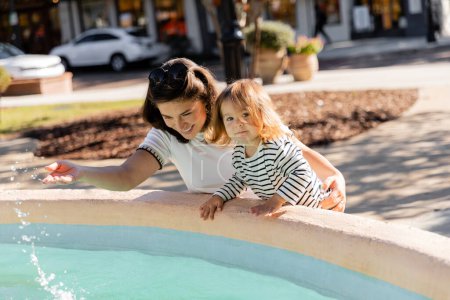glückliche Mutter blickt auf Wasser in Brunnen neben Kleinkind Tochter auf Straße in Miami 