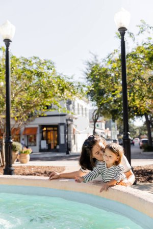 mother kissing cheek of pleased toddler daughter near fountain on street in Miami 