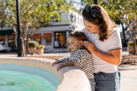Glückliche Mutter mit Sonnenbrille blickt auf süße Kleinkind-Tochter in der Nähe eines Brunnens in Miami 