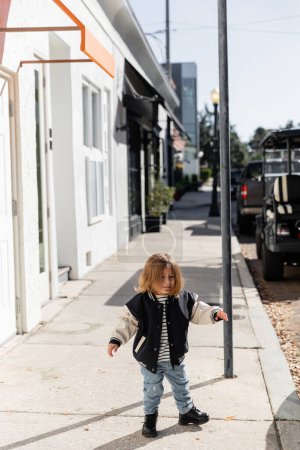 full length of happy baby girl in stylish bomber jacket walking on street in Miami 
