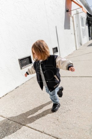 full length of baby girl in stylish bomber jacket walking on street in Miami 