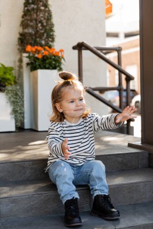 Kleinkind in lässiger Kleidung fordert Aufmerksamkeit und gestikuliert, während es auf einer Treppe in der Nähe des Hauses sitzt 