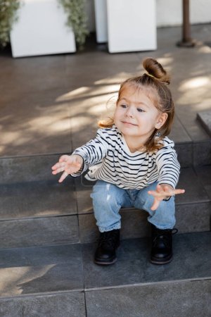 baby girl in casual clothes demanding attention and gesturing while sitting on stairs near house 