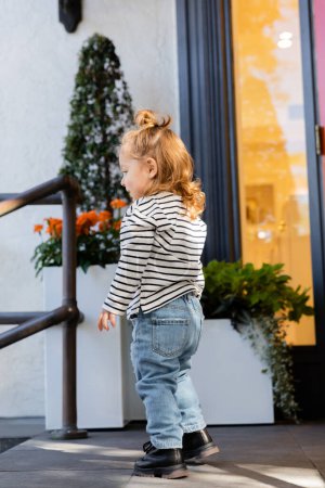 Téléchargez les photos : Vue latérale de bébé fille en chemise à manches longues et jeans bleus debout près de la maison à Miami - en image libre de droit