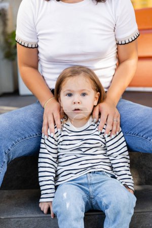 mother in jeans sitting with amazed toddler daughter on porch at backyard against house 