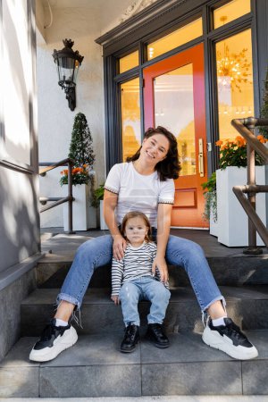glückliche Mutter in Jeans sitzt mit Kleinkind Tochter auf Veranda im Hinterhof gegen Haus in Miami 