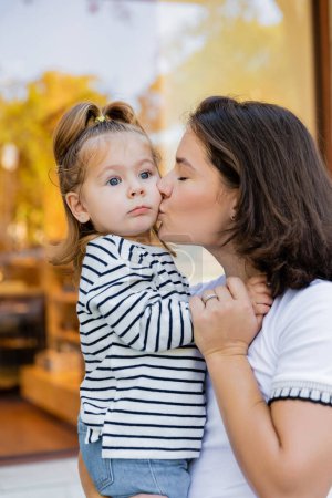 Photo pour Mère brune embrasser bébé fille à rayures chemise à manches longues près vitrine de magasin à Miami - image libre de droit