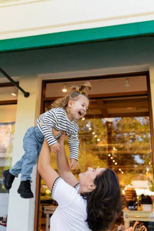 cheerful mother lifting happy baby girl near showcase of shop in Miami 