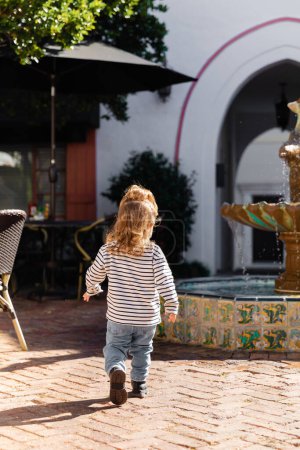 Téléchargez les photos : Vue arrière de bébé fille tout-petit en chemise à manches longues rayée et jeans marchant près de la fontaine à Miami - en image libre de droit