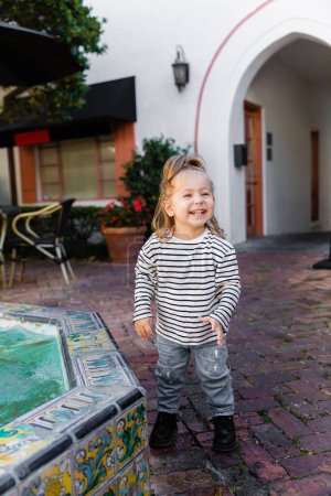 Foto de Niña feliz en camisa de manga larga a rayas de pie cerca de la fuente en Miami - Imagen libre de derechos