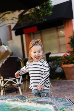 Téléchargez les photos : Joyeuse petite fille en chemise à manches longues rayée debout près de la fontaine à Miami - en image libre de droit