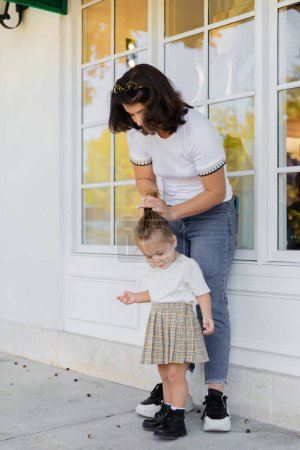 Téléchargez les photos : Mère en jeans faire des cheveux de fille tout-petit en jupe sur le porche de la maison - en image libre de droit