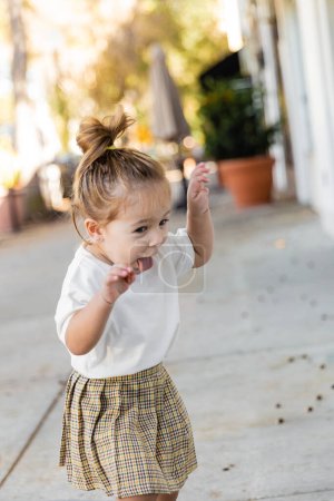 toddler girl in skirt and t-shirt sticking out tongue and gesturing outdoors 