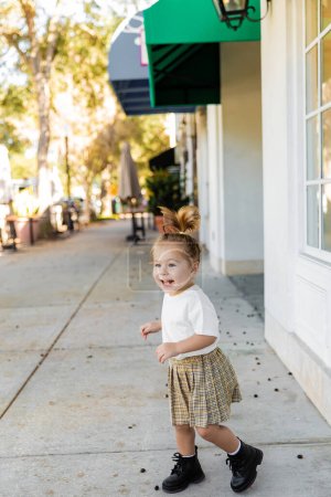 full length of positive toddler girl in skirt and t-shirt standing near building 