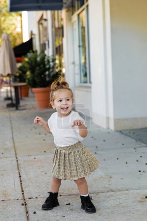 full length of positive toddler girl in skirt and white t-shirt standing on street in Miami 