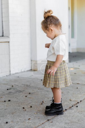 full length of toddler girl in skirt and white t-shirt looking at acorns on ground 