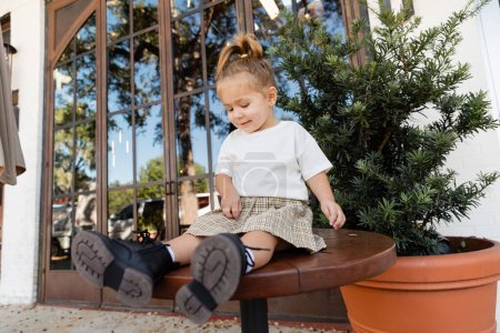 full length of smiling toddler girl in skirt and white t-shirt sitting on bistro table near outdoor cafe in Miami 