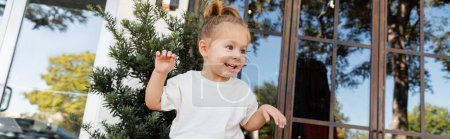 cheerful toddler girl in white t-shirt smiling near outdoor cafe on street in Miami, banner 