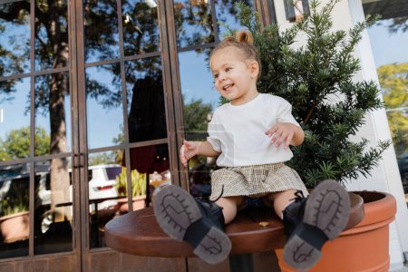 joyful toddler girl in skirt and white t-shirt sitting on wooden bistro table near outdoor cafe in Miami 