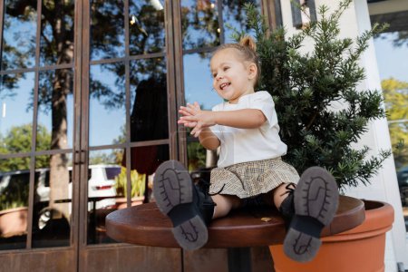 positive toddler girl in skirt and white t-shirt clapping hands while sitting on bistro table in Miami 