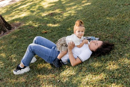 brunette mother pouting lips and lying on grass with toddler baby girl 