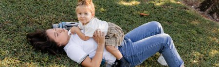 brunette mother pouting lips and lying on grass with toddler baby girl, banner 