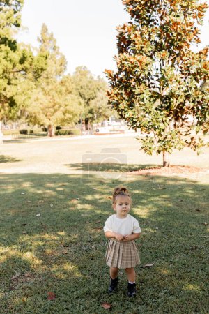 full length of toddler girl in skirt and white t-shirt standing in park with green trees 