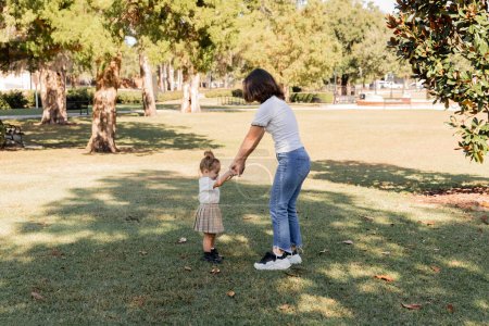 full length of mother in jeans holding hands with toddler daughter in park of Miami 